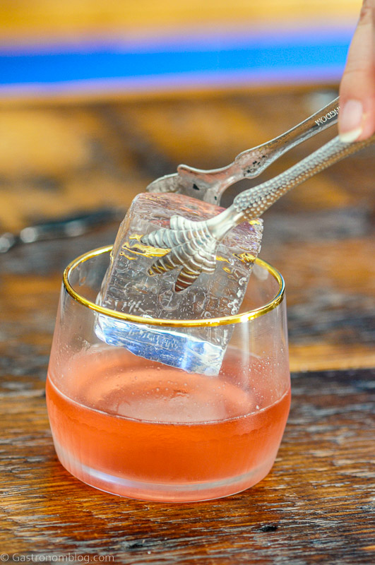 Ice cube being placed in gold rimmed glass with talon ice tongs