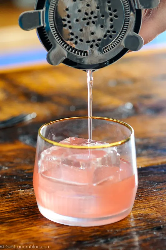 Pink cocktail being poured from cocktail shaker into gold rimmed glass with clear ice