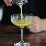 Yellow cocktail being poured into cocktail coupe