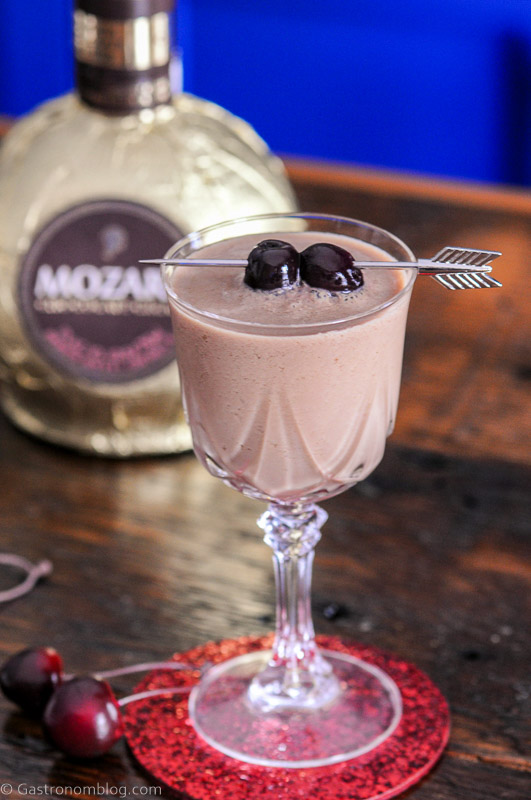 Chocolate cocktail in coupe with cherries on arrow pick, liqueur bottle behind