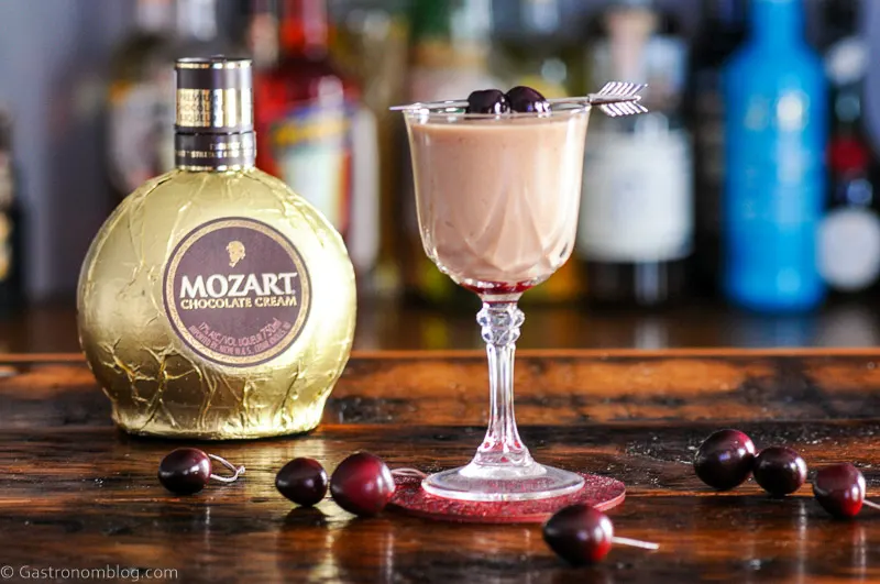 Chocolate cocktail in coupe with cherries on arrow pick, liqueur bottle behind