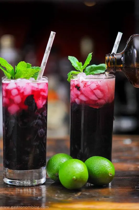 Blueberry Non Alcoholic Mojito, dark red cocktails in tall glasses, limes next to glasses. Mint sprigs and silver dotted straw