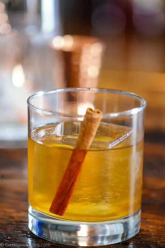 cocktail with cinnamon stick and clear ice in rocks glass, mixing glass behind