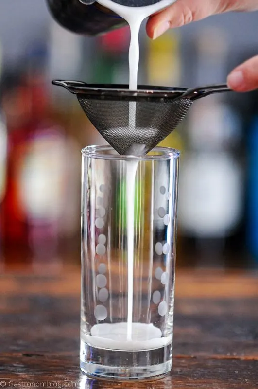 Cocktail being poured into tall glass from shaker through a mesh strainer