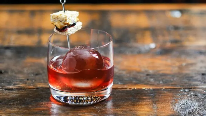 Pink cocktail in rocks glass with clear ice ball, tiny peanut butter and jelly sandwich