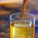 Brown cocktail in rocks glass with ice, cinnamon stick being placed in