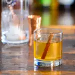cocktail with cinnamon stick and clear ice in rocks glass, mixing glass behind