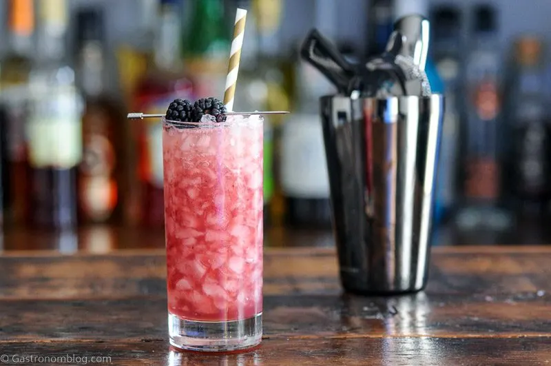 Purple cocktail in tall glass with crushed ice, blackberries on pick and gold and white straw