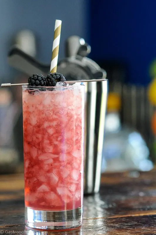 Purple whiskey collins cocktail in tall glass with crushed ice, blackberries and gold/white straw