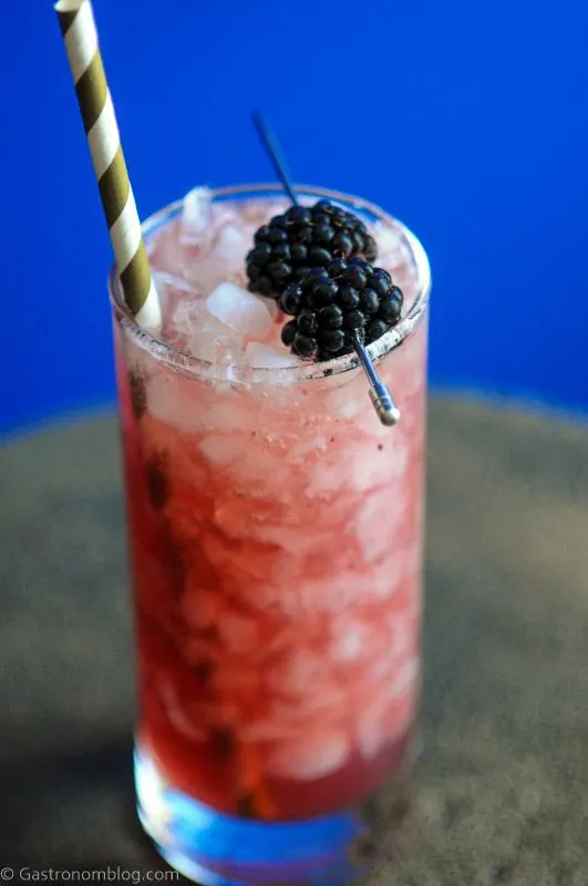 Purple whiskey collins cocktail in tall glass with crushed ice, blackberries and gold/white straw