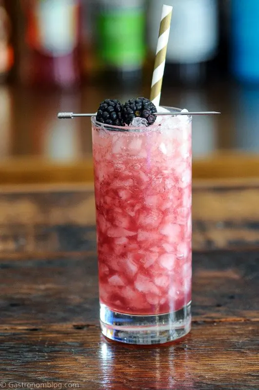 Purple cocktail in tall glass with crushed ice, blackberries on pick and gold and white straw