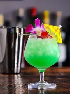 Blue green cocktail in balloon glass with crushed ice, paper umbrella, cherry and orchids