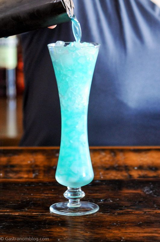 Blue cocktail being poured into tall glass from cocktail shaker