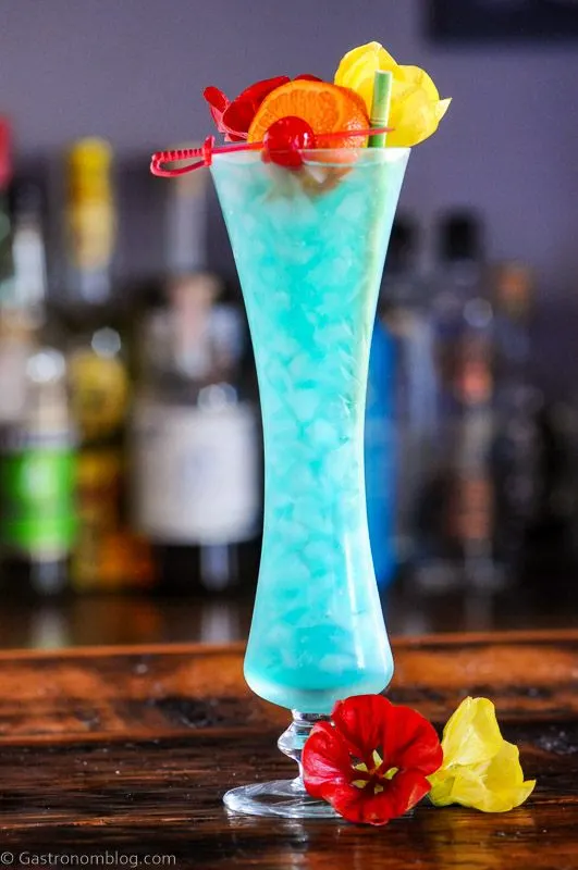 Blue Lagoon Cocktail recipe in a tall glass with flowers, cherry on a pick and an orange slice