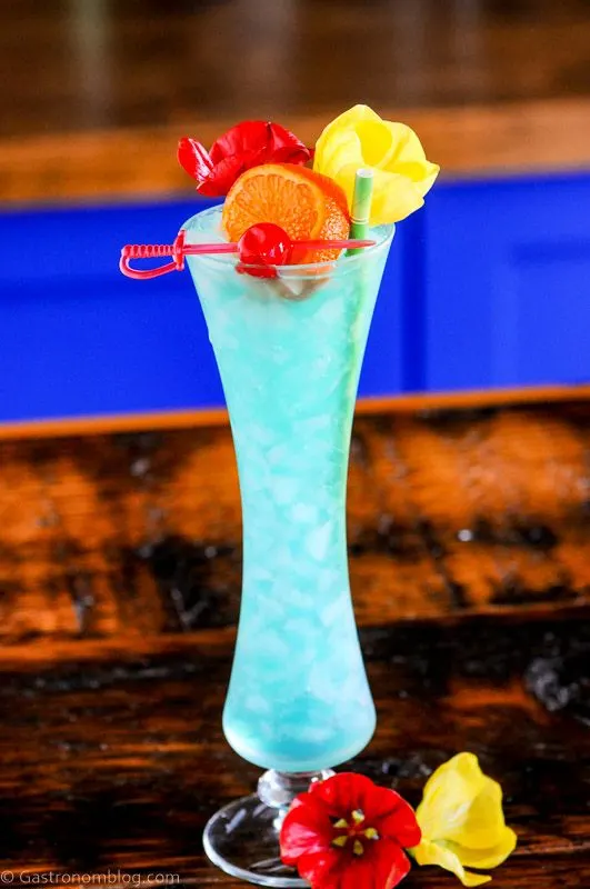 Blue Lagoon Cocktail recipe in a tall glass with flowers, cherry on a pick and an orange slice