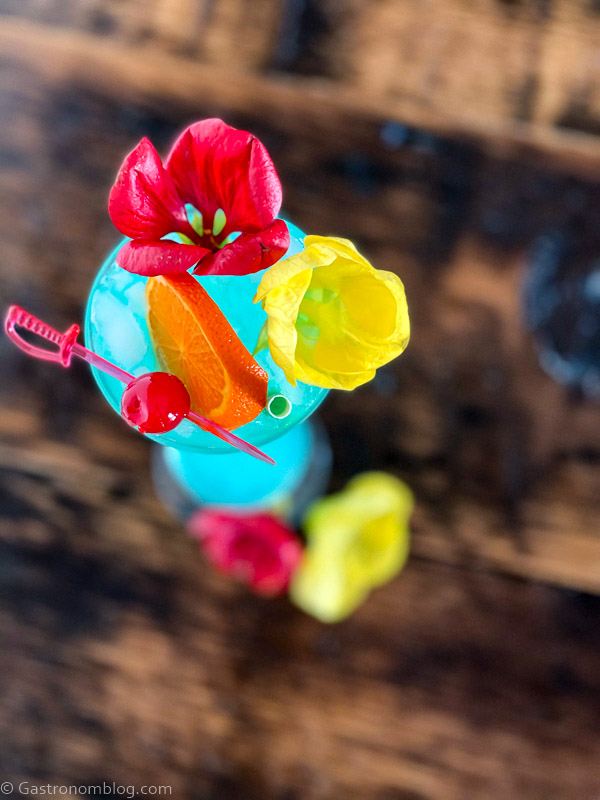Top shot of a blue cocktail, flowers, cherry on a pick and an orange slice