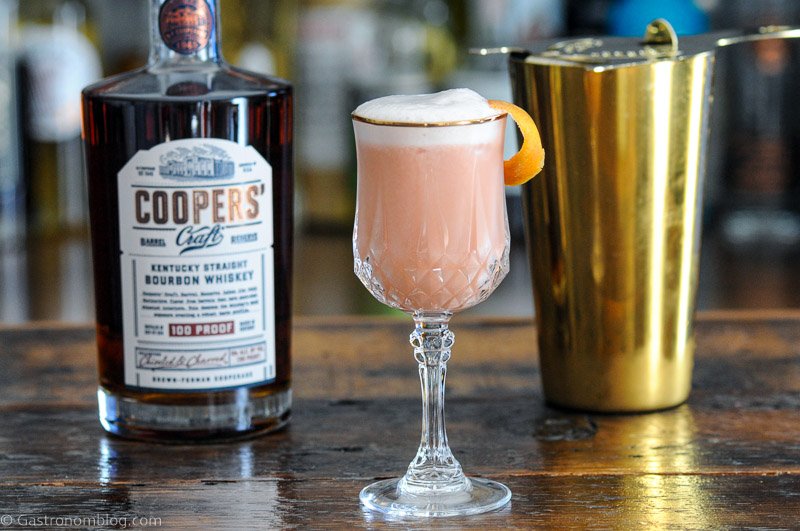 Pink cocktail with white foam in gold rimmed glass. Whiskey bottle behind