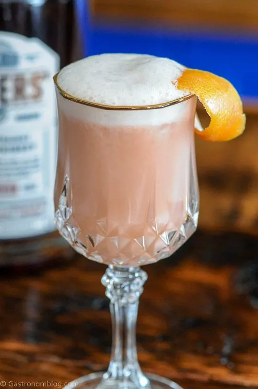 Pink cocktail with white foam in gold rimmed glass. Whiskey bottle behind