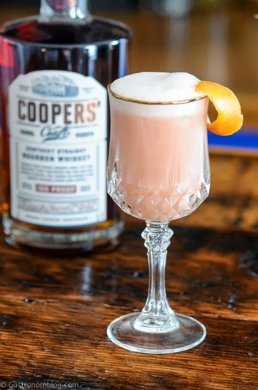 Pink cocktail with white foam in gold rimmed glass. Whiskey bottle behind