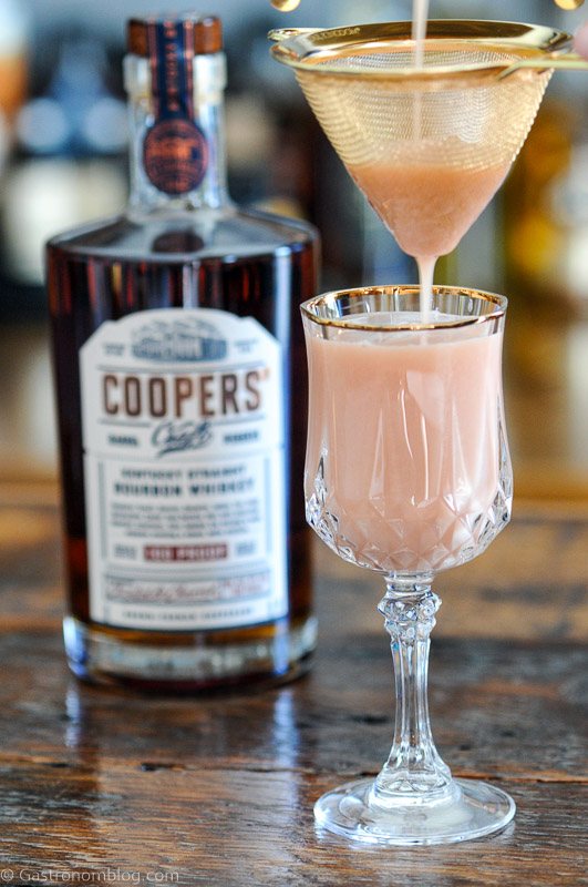 Pink cocktail being strained into glass from cocktail shaker, Coopers' Craft bottle behind