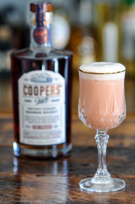 Grapefruit Bourbon Sour, a pink cocktail in glass with white foam topping. Whiskey bottle behind