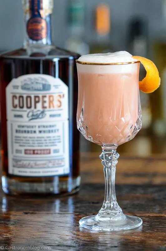 Pink cocktail with white foam in gold rimmed glass. Whiskey bottle behind