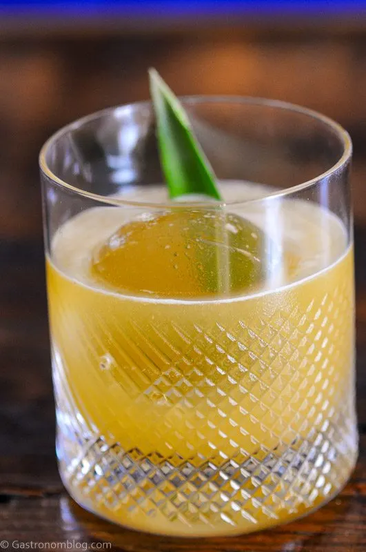 Yellow cocktail in rocks glass with clear ice and pineapple frond