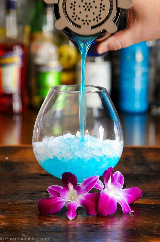 Blue Curacao cocktail being poured into bulb glass, purple and white orchids