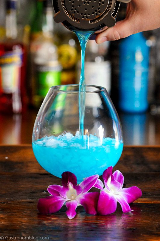 Blue Curacao cocktail being poured into bulb glass, purple and white orchids