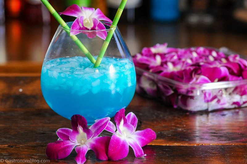 Blue Curacao cocktail being poured into bulb glass, purple and white orchids, bamboo straws