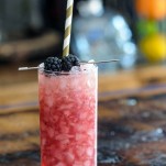 Purple whiskey collins cocktail in tall glass with crushed ice, blackberries and gold/white straw