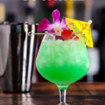 Green cocktail in bulb glass, purple orchid, cherry and yellow umbrella, shaker in background