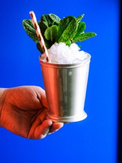 hand holding silver cup with crushed ice, mint sprig and striped paper straw in front of a blue cabinet