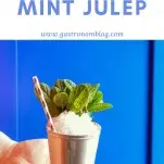 Hand holding silver julep cup piled with crushed ice, mint sprig and striped paper straw in front of blue cabinet
