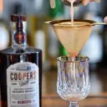 Pink cocktail being strained into glass from cocktail shaker, Coopers' Craft bottle behind