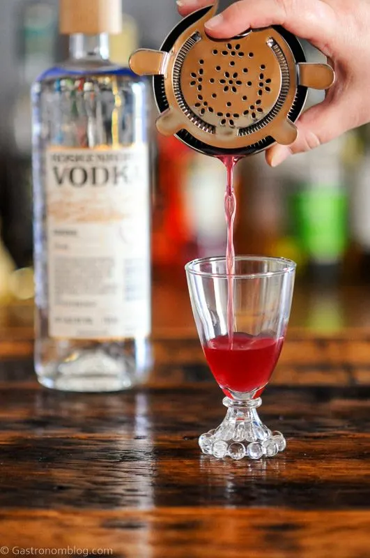 Pink cocktail being poured into glass from a shaker with a hawthorne strainer, vodka bottle in background