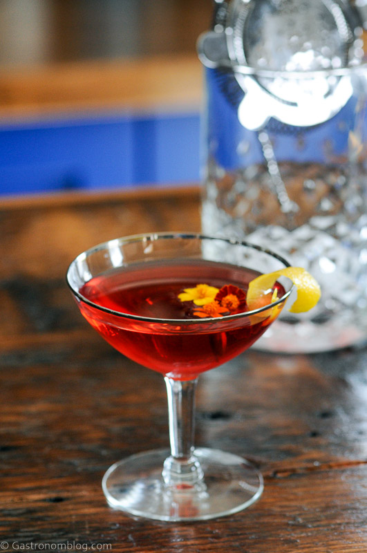 Red cocktail in coupe with lemon peel on edge, mixing glass behind
