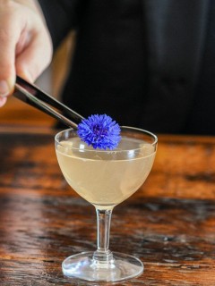 opaque cocktail in coupe with purple flower being placed by tweezers