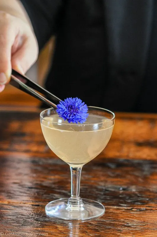 opaque cocktail in coupe with purple flower being placed by tweezers