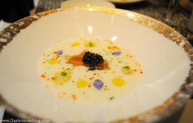 caviar on top of white soup with colored oils and edible flowers in gold rimmed bowl