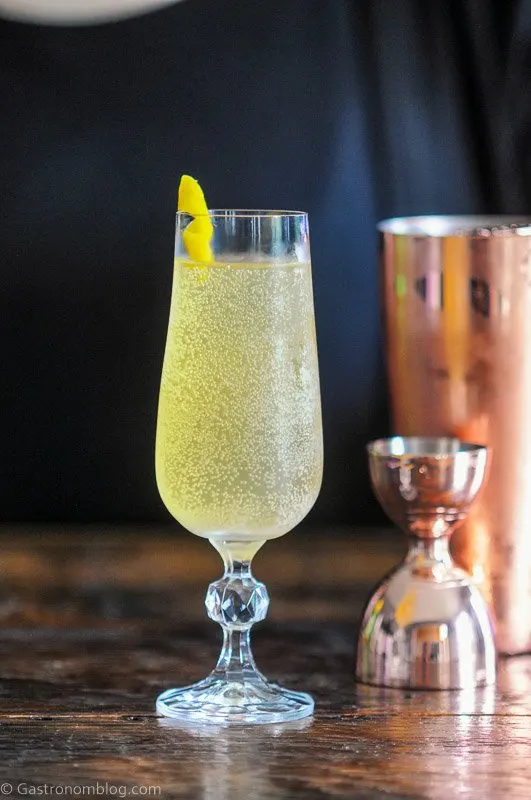 gold cocktail, Cognac French 75, in tall glass, lemon peel garnish, gold cocktailware behind