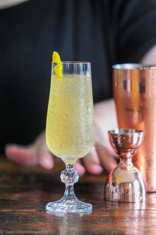 gold cocktail, Cognac French 75, in tall glass, lemon peel garnish, gold cocktailware behind