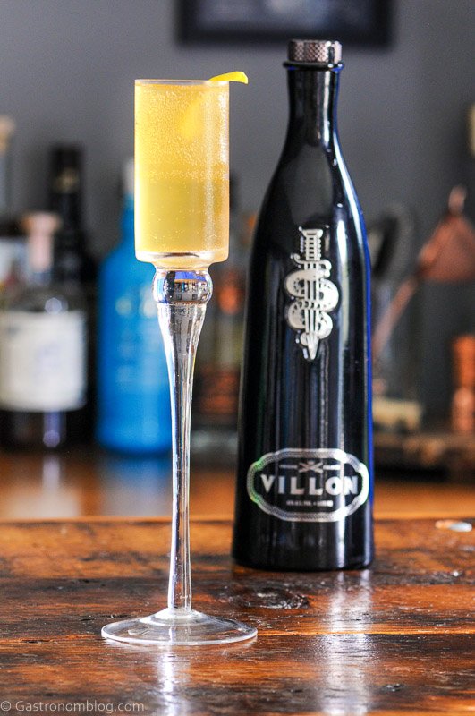 Villon Cognac Liqueur French 75, Gold cocktail in tall glass, Black glass bottle behind