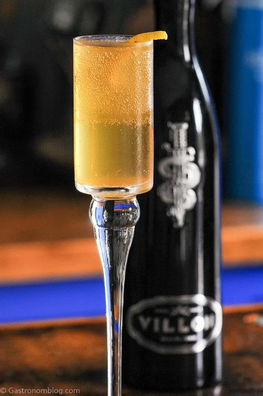 Villon Cognac Liqueur French 75, Gold cocktail in tall glass, Black glass bottle behind