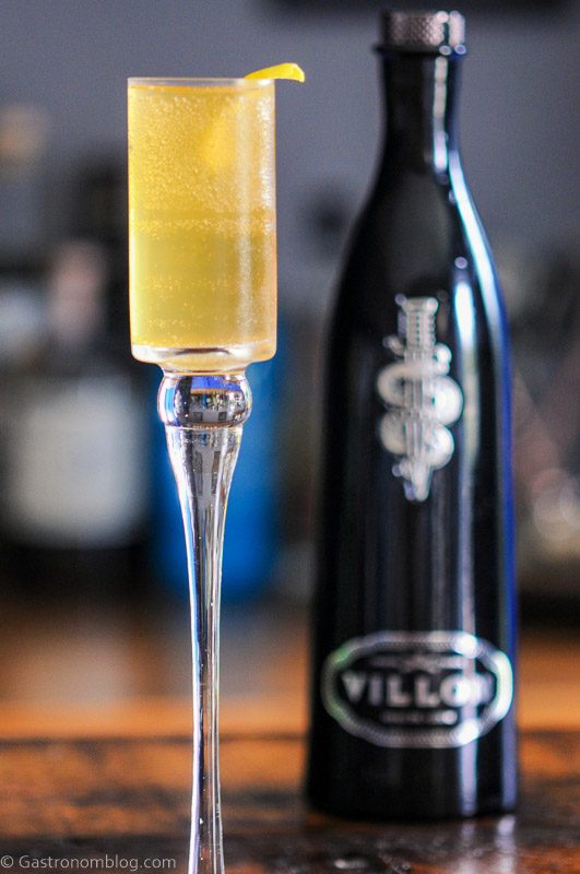 Villon Cognac Liqueur French 75, Gold cocktail in tall glass, Black glass bottle behind
