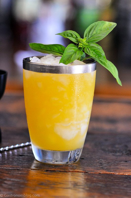 Orange cocktail in silver rimmed glass, basil leaves on top, black jigger