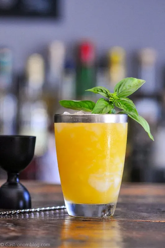 Orange cocktail in silver rimmed glass, basil leaves on top, black jigger