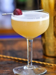Yellow cocktail with white foam in cocktail coupe, cherry on cocktail pick. Gold barware behind