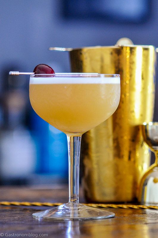 Peach Bourbon Sour, yellow cocktail with white foam in cocktail coupe, cherry on pick. Gold Barware behind