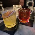 Yellow cocktail with grapefruit slice and brown cocktail with cherries in rocks glasses on marble table
